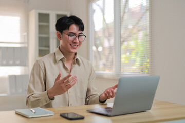 Young Asian man smiling and gesturing during a casual remote video call from his home office, laptop and webcam visible, professional yet relaxed
