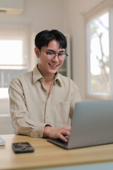 Young Asian man wearing glasses smiling and typing on laptop while sitting at a desk in a home office