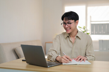 Smiling young man wearing glasses using a laptop and writing in a notebook, focused on remote work or education