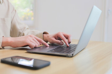 Person's hands working on a laptop computer with a smartphone on a wooden desk, representing remote work and technology use
