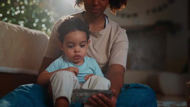 Be happy in new year and celebrate merry Christmas, african mom and son at home. Careful mother holding cute toddler and showing smartphone with cartoons, calm atmosphere of cozy home, winter holidays