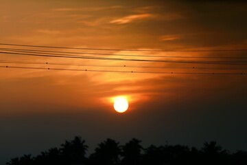 Fototapeta premium Dramatic sunset with a bright orange sun and layered clouds above a dark rural treeline, capturing calm and natural beauty