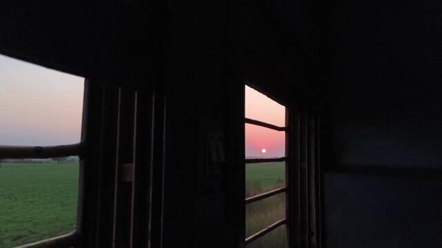 Hubli, Karnataka, India - December 13, 2025: The sun sets over green fields, framed by metal bars of a moving train window, creating a quiet and reflective travel moment.
