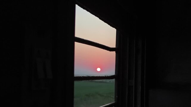 Hubli, Karnataka, India - December 13, 2025: The sun sets over green fields, framed by metal bars of a moving train window, creating a quiet and reflective travel moment.
