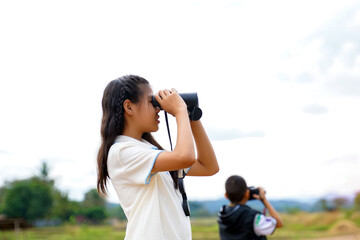 A young girl focused on observing nature through black binoculars, standing outdoors in a green field or park during a daytime exploration trip.