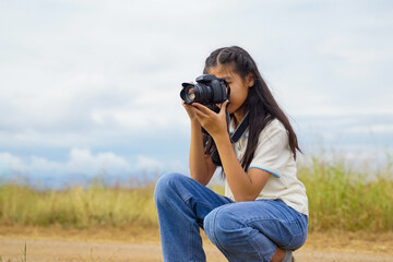 A young girl holding a digital camera and looking through the viewfinder, capturing a photo of the surrounding nature in a park or forest.