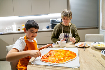 Grandmother and grandson bonding while preparing homemade pizza for dinner, spreading tomato sauce on dough