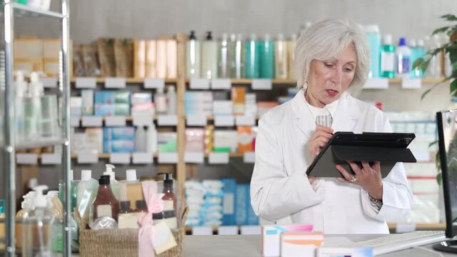 Mature female pharmacist works with a stylus on a tablet, she counts the leftovers of drugs in the pharmacy warehouse and plans a restock. Pharmacist works in a pharmacy. High quality 4k footage