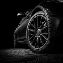 Sleek, dark car close-up, with the wheel prominent on rough ground, lit in chiaroscuro against a black backdrop