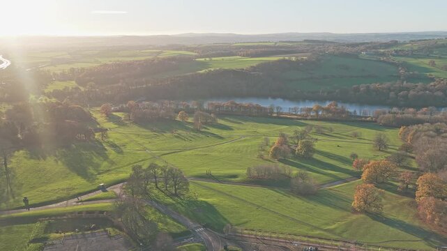 Drone flyover capturing landscape of the Yorkshire Sculpture Park, features large ornamental Bretton Lake, manicured green fields, and paths, all dramatically lit by the late afternoon sun, England.