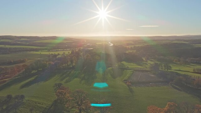 Slow drone push-in shot, centered on a powerful, vertical sunburst captures the expansive, undulating green parkland of the Yorkshire Sculpture Park and its scattered trees in autumn season