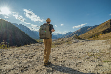 A solo hiker in bright clothing walks a mountain road in the Altai on a sunny day. Rear view captures the trekker against a backdrop of snowy peaks and autumn forest.