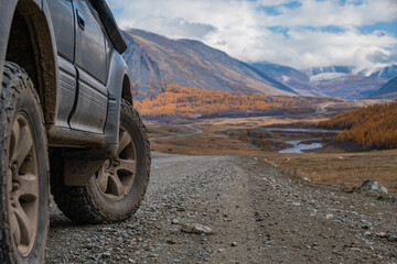 Off-road vehicle tires captured in medium shot on a mountain dirt road slope. The backdrop features vibrant autumn forest, a flowing mountain river, and towering peaks.