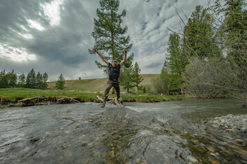 A trail runner in waterproof boots splashes through a small mountain river in the Altai forest. Action shot captures the spray against a backdrop of green hills and conifers.