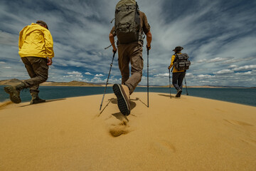 A group of friends with trekking poles and backpacks hikes up a massive sand dune in the Gobi Desert. Low rear-angle shot captures them against an emerald lake and vast dunes.