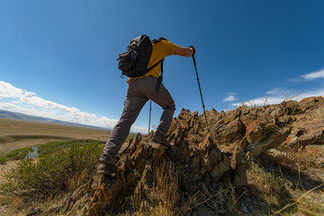 A hiker with backpack and trekking poles ascends a rocky hill with sharp ridges. Medium side shot captures the effort against a vast Mongolian steppe and blue sky.