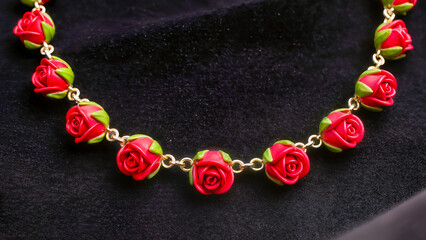 An elegant close-up of a jewelry piece or high-quality costume jewelry. A delicate golden chain is adorned with a row of detailed charms in the shape of blooming red roses with green sepals. The piece