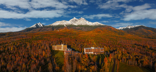 Tatranske Matliare, Slovakia - Aerial view of snowy mountains of Lomnicky Peak in the High Tatras with hotels, beautiful red and orange coloured autumn trees and foliage and blue sky at Vysoke Tatry