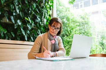 Mature woman working at a cafe with a laptop and financial documents. She reviews papers intently...