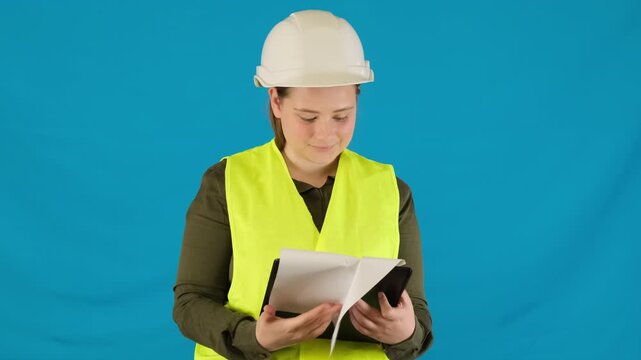Positive female engineer examines papers holding clipboard on blue background. Woman in helmet and work vest checks project documentation