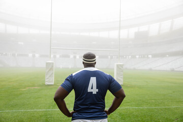 Rugby player is standing on well-kept grass pitch facing rugby goalposts amid light fog