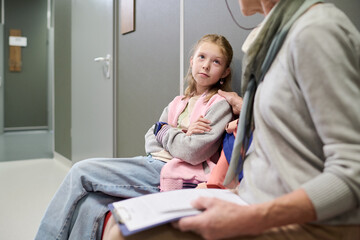 Obraz premium Caucasian girl sitting with arms crossed next to senior Caucasian woman holding clipboard, both waiting in hallway, girl looking up attentively at woman, casual setting