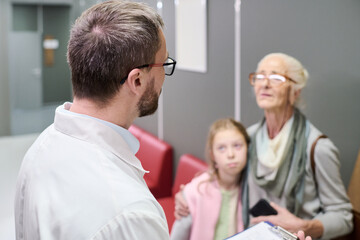 Obraz premium Caucasian male doctor speaking with senior Caucasian woman and young Caucasian girl in medical office, senior woman holding smartphone while girl standing close to her