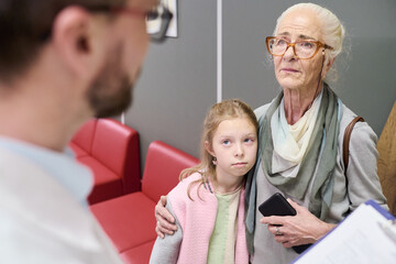 Obraz premium Senior Caucasian woman standing with arm around young Caucasian girl while listening to male doctor, both looking concerned, girl holding smartphone, indoor setting