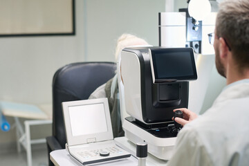 Caucasian senior woman undergoing eye examination with autorefractor operated by man in medical...
