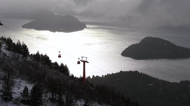 Snowy Cerro Otto cable car tower above shimmering lake and forested islands under dramatic winter sky.