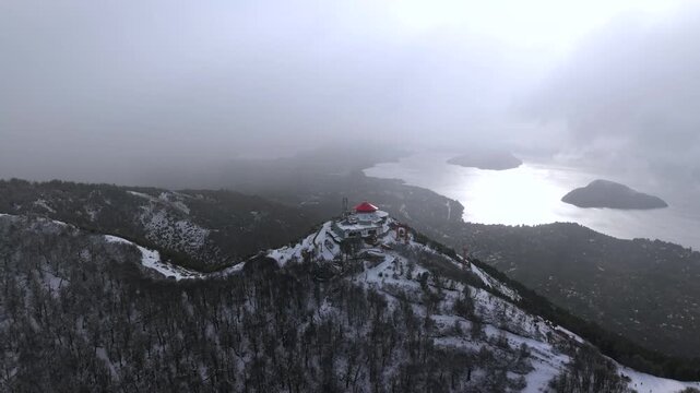 Snowy Cerro Otto ridgeline leading to red-roof summit building above Bariloche lakes under stormy sky.