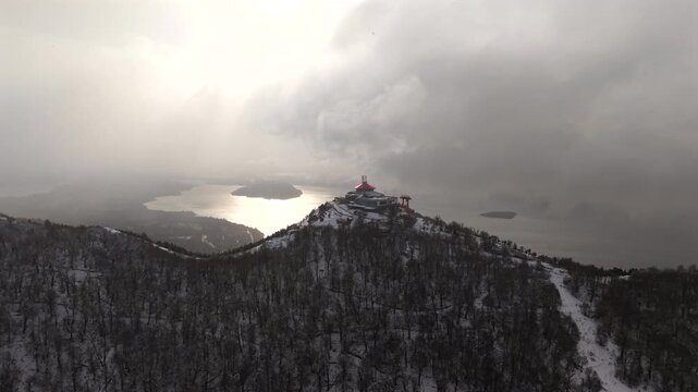 Snowy Cerro Otto ridgeline with red-roof summit building and winding road above Bariloche lakes at dusk.