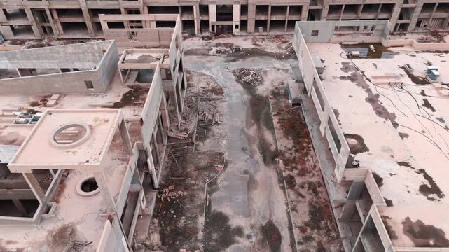 Aerial drone view of a large abandoned construction site. Unfinished concrete buildings with overgrown weeds and debris, showing urban decay and halted development.