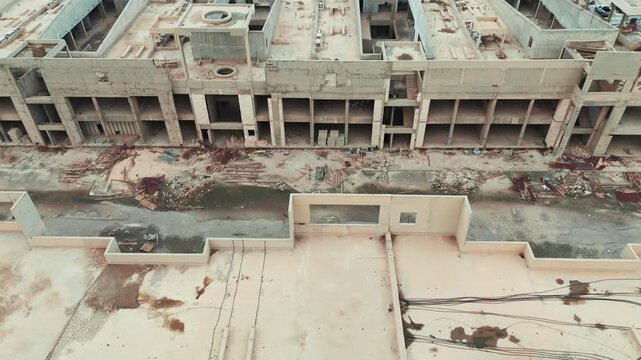 Aerial drone view of a massive, unfinished commercial building complex. Decaying concrete skeleton with unused materials and weeds, symbolizing economic recession and halted development.