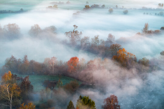 Aerial view of ethereal mists dance across the landscape, veiling the trees in a soft, dreamlike embrace as the river meanders through the scene, Thames Valley, Oxfordshire, United Kingdom.
