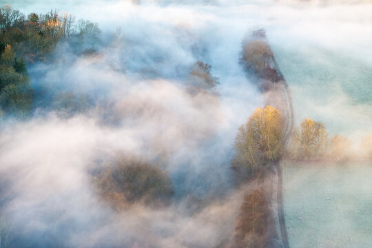Aerial view of ethereal mist blankets the landscape, partially veiling the trees and fields in soft, diffused light, Thames Valley, Oxfordshire, United Kingdom.