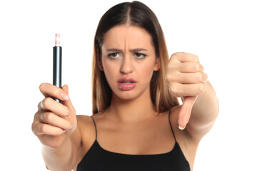 Obraz premium Studio shot of a woman with long brown hair and makeup wearing a black top, holding an electronic cigarette forward with a displeased expression and showing a thumbs down gesture on a white background
