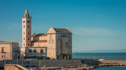 Fototapeta premium Sunlit stone cathedral with a tall bell tower in Trani, Italy, stands by the Adriatic Sea under a clear blue sky. A wide piazza, a few pedestrians, and historic buildings frame the scene.