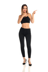 Full length studio shot of a tall brunette woman with long hair wearing a black crop top, skinny jeans and high heels pointing sideways as if indicating direction or text on a white background