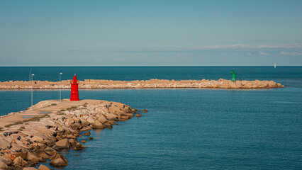 Red and green lighthouses mark the harbor entrance in Trani, Italy, set on stone breakwaters surrounded by calm blue Adriatic Sea under a clear sky, creating a peaceful coastal scene.