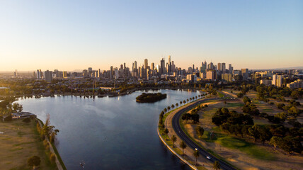 Aerial view of a tranquil lake reflecting the city skyline under the warm glow of the setting sun, with a winding road tracing its edge, Albert Park, Melbourne, Victoria, Australia.