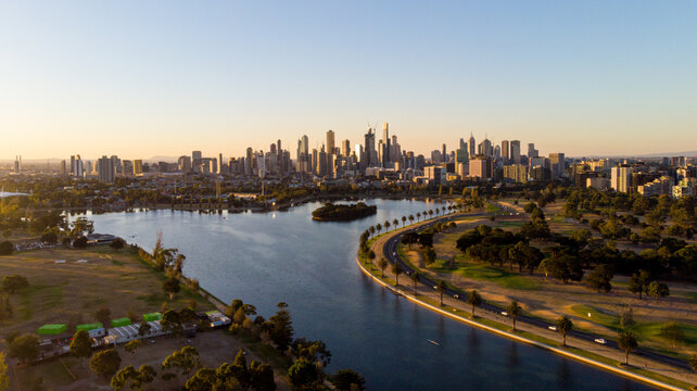 Aerial view of the glimmering Albert Park Lake reflecting the city skyline under a soft sunrise, a serene vista contrasting with the bustling cityscape, Albert Park, Victoria, Australia.