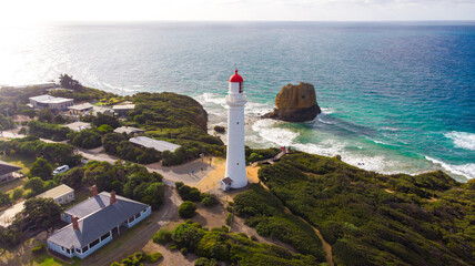 Aerial view of the iconic white Split Point Lighthouse with its striking red top standing tall against the turquoise sea and rugged coastline, Split Point Lighthouse, Victoria, Australia.