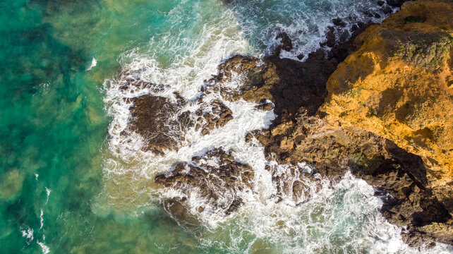 Aerial view of frothy waves crashing against the rugged, golden coastline near Eagle Rock Parade, a vibrant clash of turquoise sea and ochre cliffs, Split Point Lighthouse, Victoria, Australia.