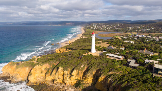 Aerial view of Split Point Lighthouse standing tall against the rugged coastline, where the turquoise sea meets the golden sands under a cloudy sky, Split Point Lighthouse, Victoria, Australia.