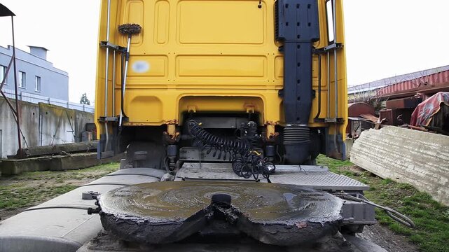 Yellow semi-truck rear. Greasy fifth wheel coupling. Air hoses, electrical connections. Heavy transport vehicle in industrial yard. Work, maintenance, daytime.