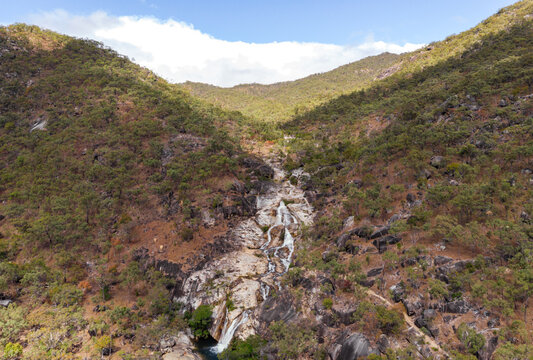 Aerial view of Emerald Falls cascading down rugged terrain, a ribbon of white against the ochre and verdant landscape, Mareeba, Queensland, Australia.