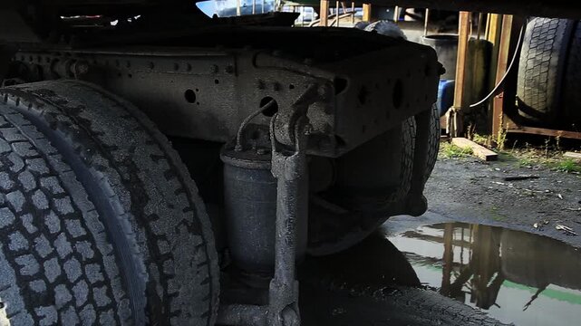 Underside view of heavy duty truck trailer. Rugged tires air suspension system robust chassis frame visible. Gritty industrial repair setting dirt grime water puddles.