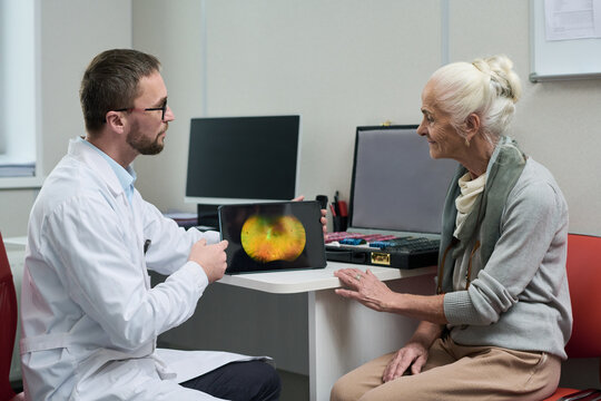 Caucasian male doctor showing digital retinal scan on tablet to senior Caucasian woman during medical consultation, both sitting at desk in clinical office, discussing eye health