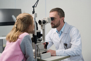 Obraz premium Caucasian middle aged man performing eye examination on Caucasian child using ophthalmology equipment in medical office, both focusing on diagnostic procedure, clinical setting visible